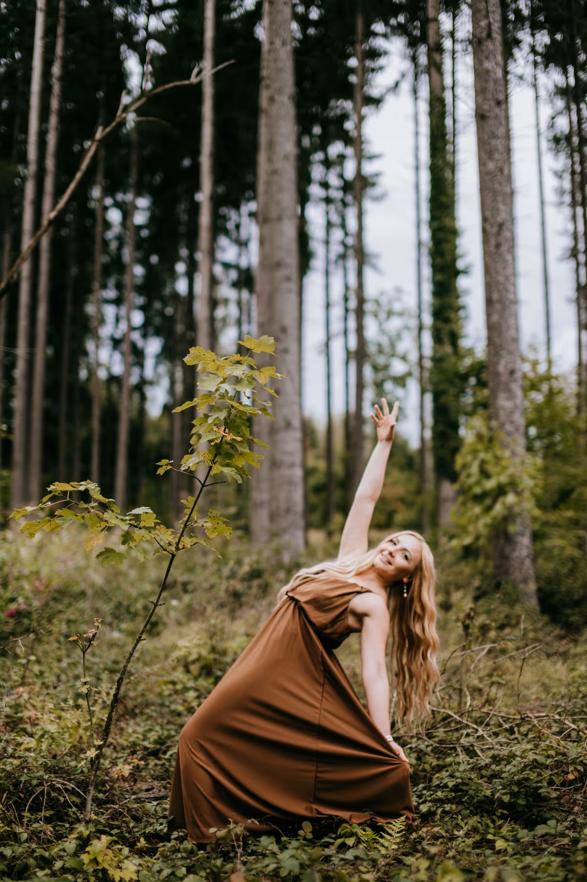 Woman in a brown dress standing in a forest with tall trees and greenery.