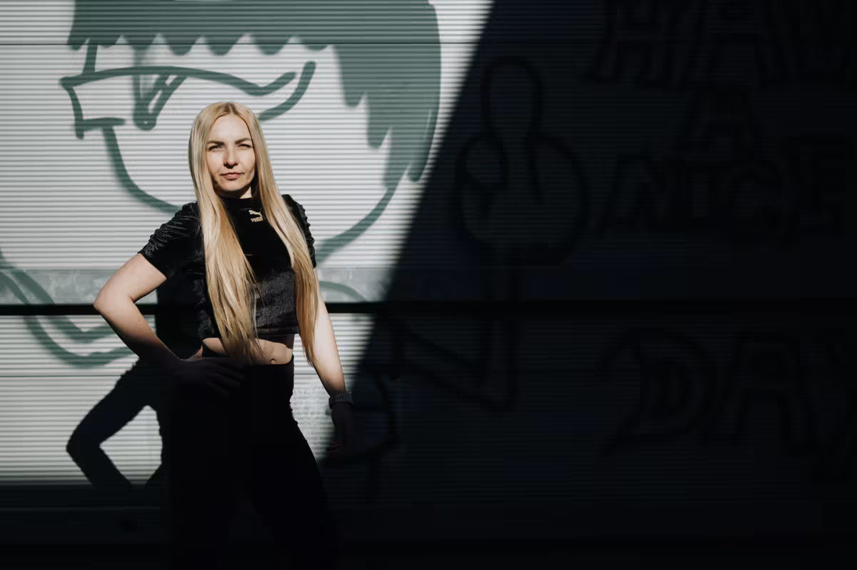 Woman in black outfit standing in front of a dark background with a logo.